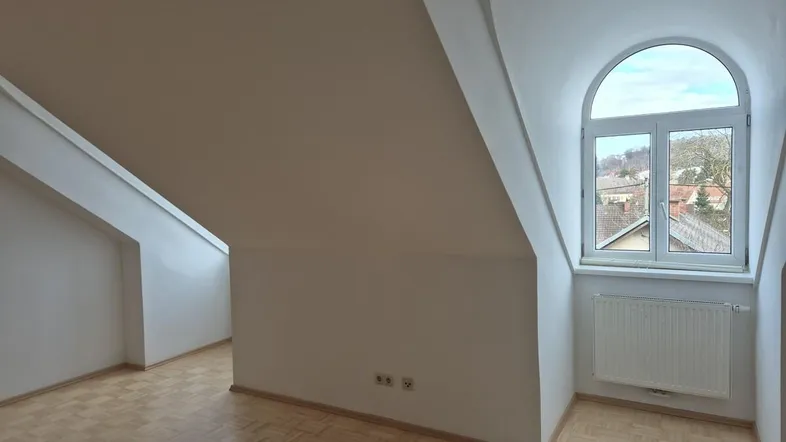 Helles Dachzimmer mit schrägen Wänden, Parkettboden und einem Bogenfenster mit Ausblick.