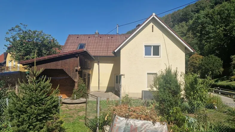 Hausfassade mit Gartenbereich und Blick auf die umliegende grüne Hügellandschaft unter blauem Himmel.