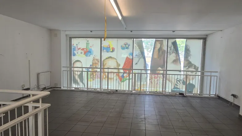 Großer, leerer Gewerberaum mit Fliesenboden, weißen Wänden und großen Fenstern mit Werbeplakaten.