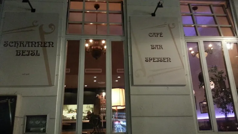 Fassade eines Cafés/Bars bei Nacht mit beleuchteten Fenstern und Beschriftungen.
