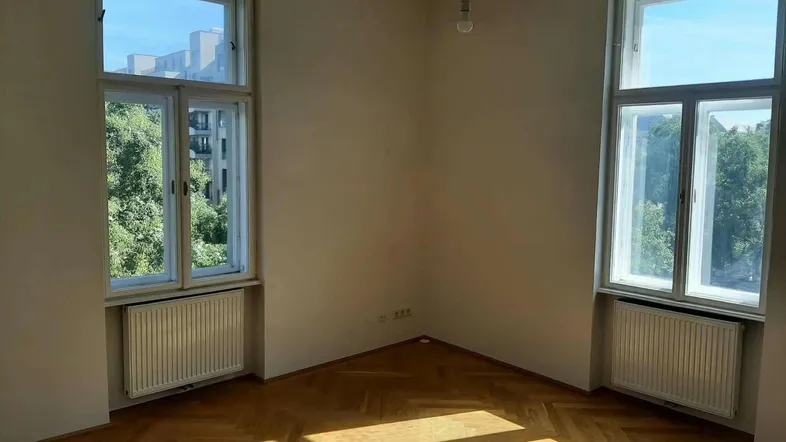 Helles Zimmer mit Fischgrätparkettboden und zwei hohen Fenstern, die viel Tageslicht hereinlassen.