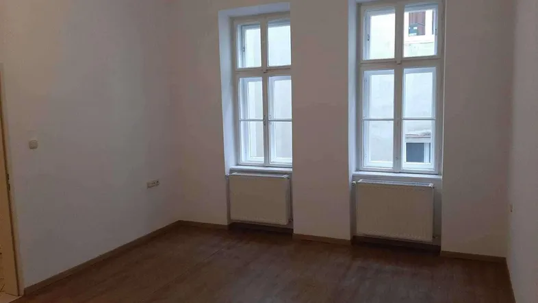 Helles, leeres Zimmer mit zwei hohen Fenstern und Heizkörpern, bereit zur individuellen Gestaltung.