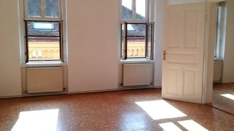 Helles Zimmer mit Parkettboden und zwei Fenstern, das viel Tageslicht hereinlässt.