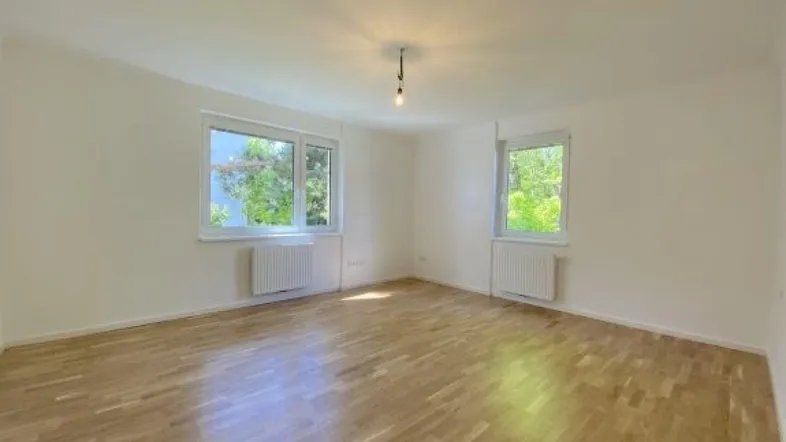 Geräumiges Zimmer mit hellem Parkettboden und Ausblick auf die umliegende Natur durch die Fenster.