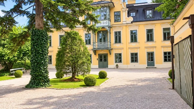 Eindrucksvolle gelbe Villa mit grünen Fensterläden und einem weitläufigen Kiesweg, umgeben von gepflegten Bäumen.