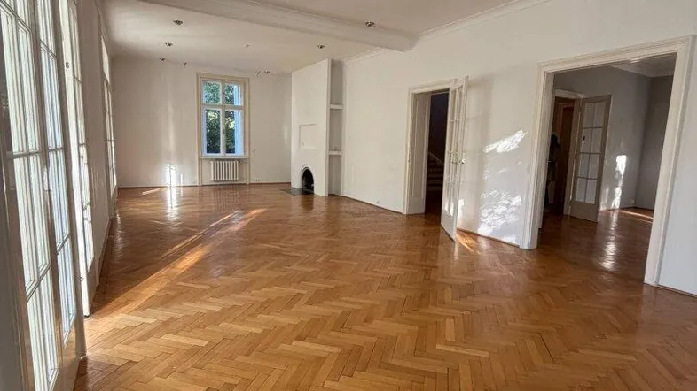 Geräumiges Wohnzimmer mit Fischgrätparkett, hohen Fenstern und einem dekorativen Kamin.