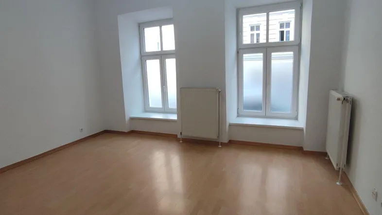 Helles Zimmer mit Parkettboden, weißen Wänden und zwei Fenstern, bereit zur individuellen Gestaltung.