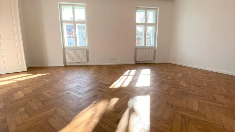 Helles, leeres Zimmer mit Fischgrätparkettboden und zwei großen Fenstern, die viel Tageslicht hereinlassen.