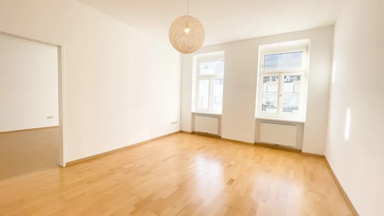 Helles, leeres Zimmer mit Parkettboden und zwei Fenstern, bereit zur individuellen Gestaltung.