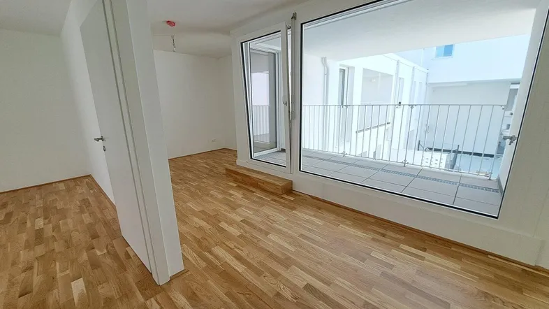 Helles Zimmer mit Holzboden und großen Fenstern, die zu einem Außenbereich führen.