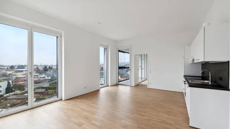 Heller offener Wohnbereich mit integrierter Küchenzeile, Parkettboden und großen Fenstern mit Stadtblick.