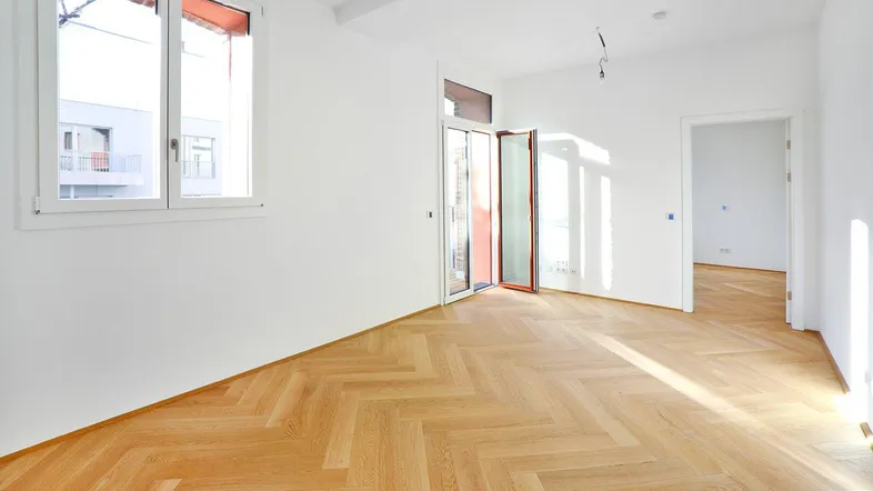 Helles, leeres Zimmer mit Fischgrätparkettboden und Zugang zu einem Balkon.