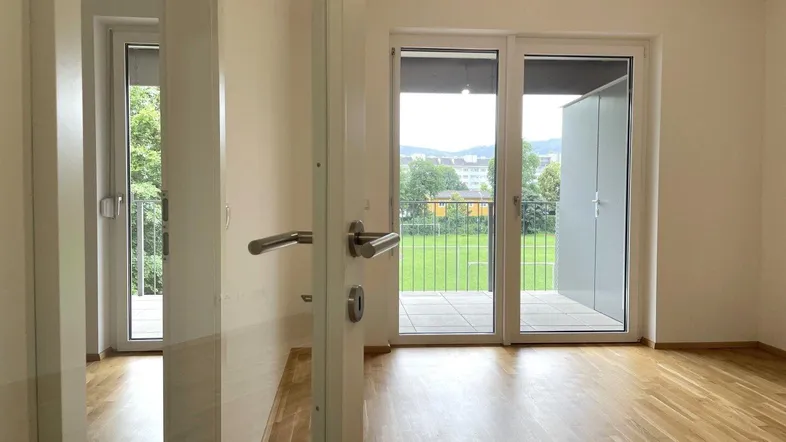 Helles Zimmer mit Parkettboden und Zugang zu einem Balkon mit Blick ins Grüne.