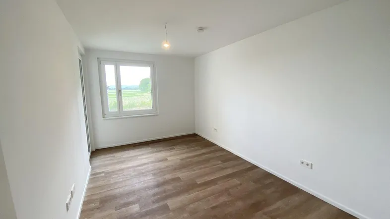 Helles, leeres Zimmer mit modernem Laminatboden und Blick ins Grüne.