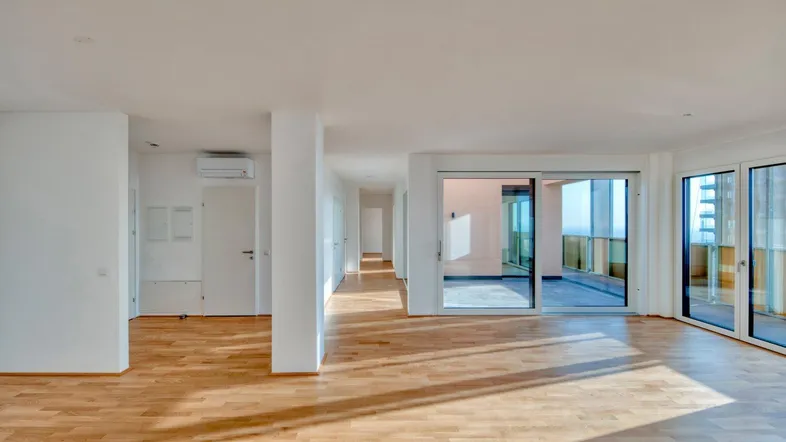 Helles, leeres Wohnzimmer mit Holzboden und großen Schiebetüren, die zu einem Außenbereich führen.