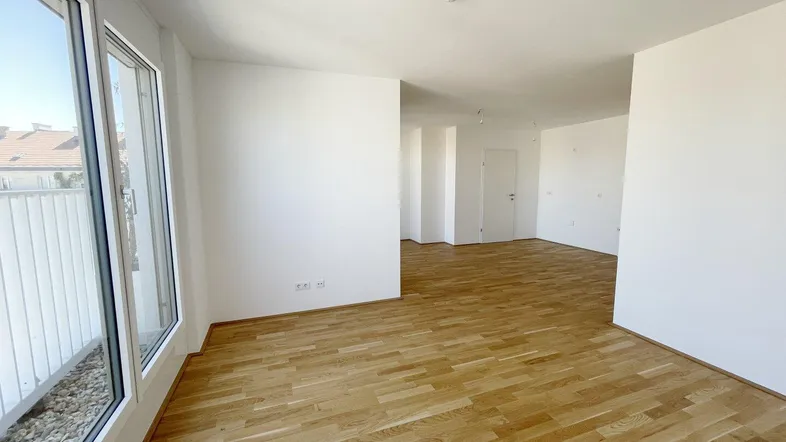 Heller, leerer Raum mit Parkettboden und großen Fenstern, die Zugang zu einem Balkon bieten.