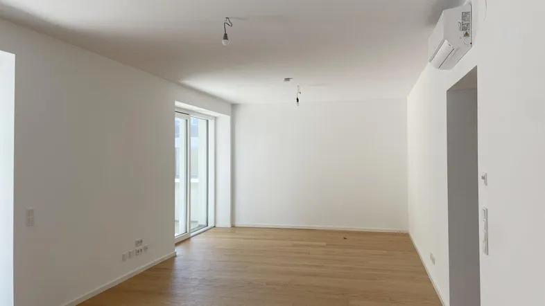 Helles, leeres Zimmer mit Holzboden und Zugang zu einem Balkon oder einer Terrasse.