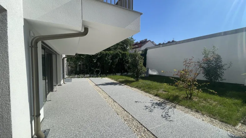 Außenbereich mit gepflastertem Weg, Rasenfläche und einem Balkon im Obergeschoss des Neubaus.