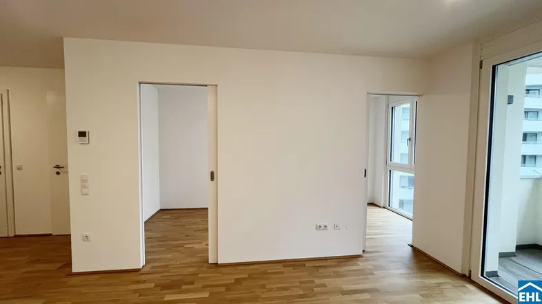 Leerer Wohnraum mit hellem Parkettboden, weißen Wänden und Zugang zum Balkon.