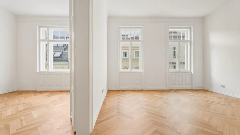 Helles Zimmer mit Fischgrätparkett und hohen Fenstern, das viel Tageslicht hereinlässt.