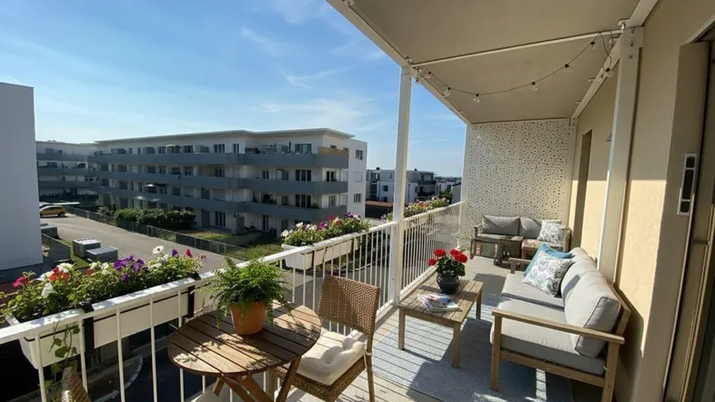 Geräumiger und sonniger Balkon mit bequemen Gartenmöbeln, Blumenkästen und Blick auf moderne Gebäude.