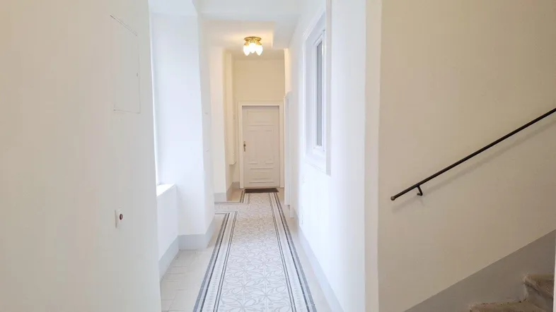 Heller Flur mit hohen Decken, Fliesenboden und einem Fenster, das Tageslicht hereinlässt.