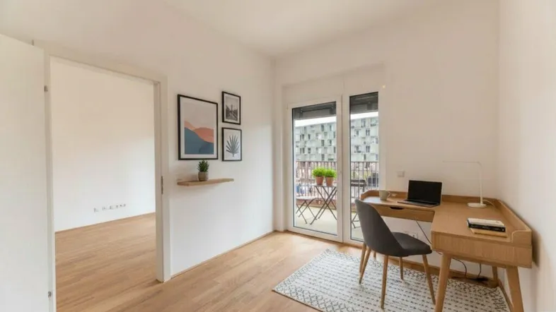 Helles Zimmer mit Holzboden, Schreibtisch und Zugang zu einem kleinen Balkon mit Stadtblick.