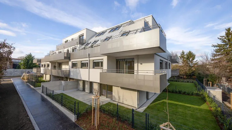 Modernes Mehrfamilienhaus mit mehreren Balkonen und Gärten, umgeben von Grünflächen unter blauem Himmel.