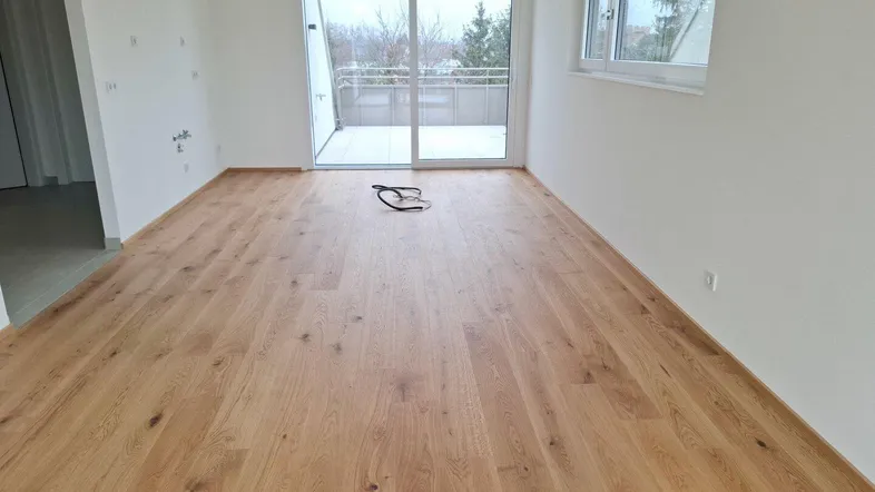 Heller, leerer Raum mit neuem Parkettboden und Zugang zu einem Balkon, bereit für die Einrichtung.