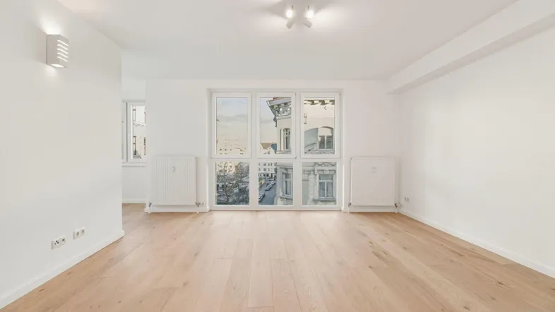Helles, leeres Zimmer mit Parkettboden und großen Fenstern, die einen Blick auf die Stadt bieten.