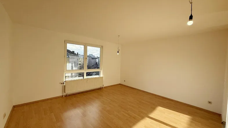 Helles, leeres Wohnzimmer mit Parkettboden und Fensterblick auf die umliegenden Gebäude.