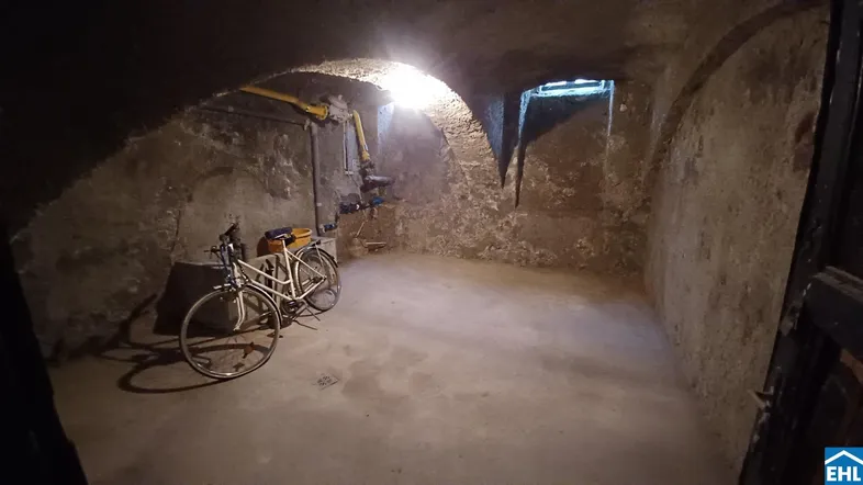 Dunkler Kellerraum mit Gewölbedecke, Betonboden und sichtbaren Leitungen, in dem Fahrräder abgestellt sind.