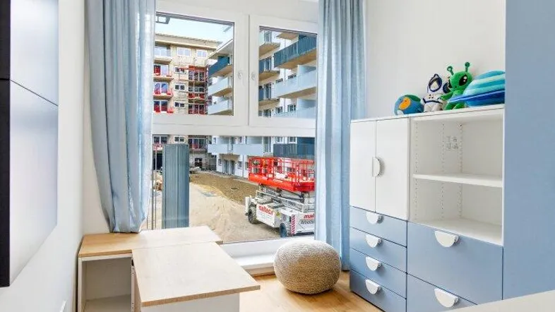 Helles Kinderzimmer mit Holzparkett, Schreibtisch und Blick auf die umliegenden Gebäude.