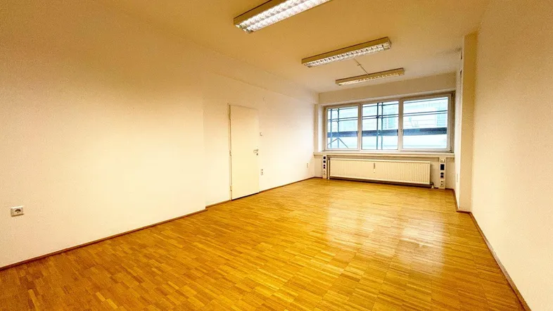Leerer Raum mit Parkettboden, großen Fenstern und Deckenleuchten.