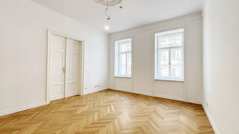 Helles Zimmer mit Fischgrätparkett, hohen Decken und Stuckverzierung an der Decke.