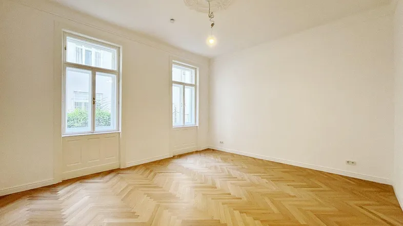 Helles Zimmer mit Fischgrätparkett, hohen Decken und Stuckverzierung an der Decke.