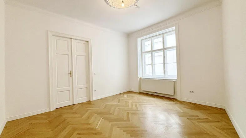 Helles Zimmer mit Fischgrätparkett, hohen Decken und einem großen Fenster, ideal als Wohn- oder Schlafzimmer.