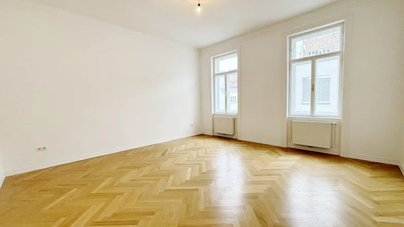 Helles Zimmer mit Fischgrätparkett, hohen Decken und Stuckverzierung an der Decke.