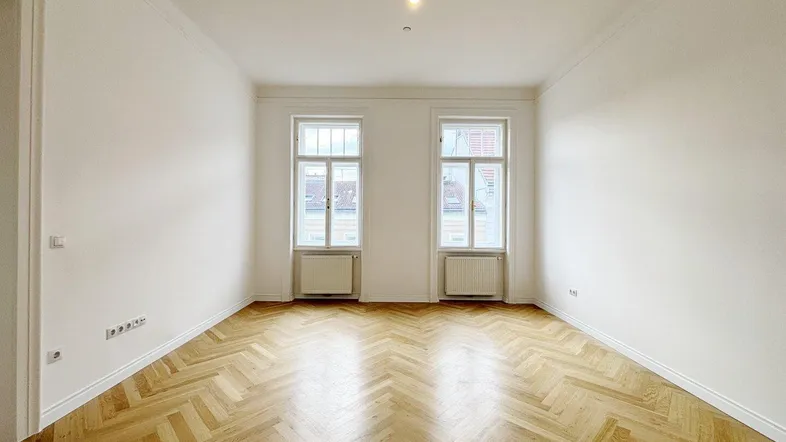 Helles Zimmer mit Fischgrätparkett, hohen Decken und dekorativem Stuck an der Decke.
