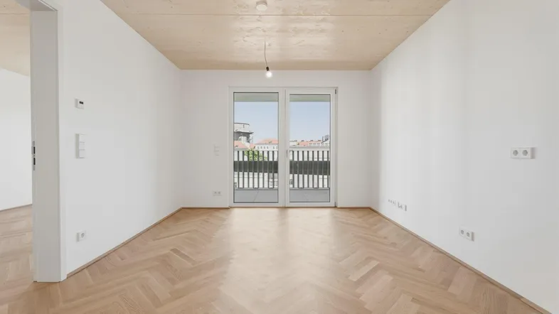 Helles, leeres Zimmer mit Fischgrätparkett und Zugang zu einem französischen Balkon.