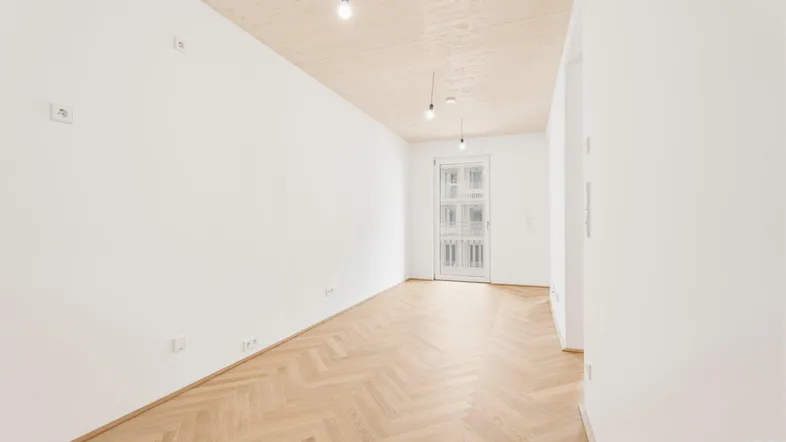 Heller, leerer Raum mit Fischgrätparkettboden, Holzbalkendecke und Zugang zu einem französischen Balkon.