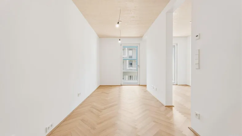 Heller, leerer Raum mit Fischgrätparkett, weißen Wänden und Zugang zu einem Balkon.