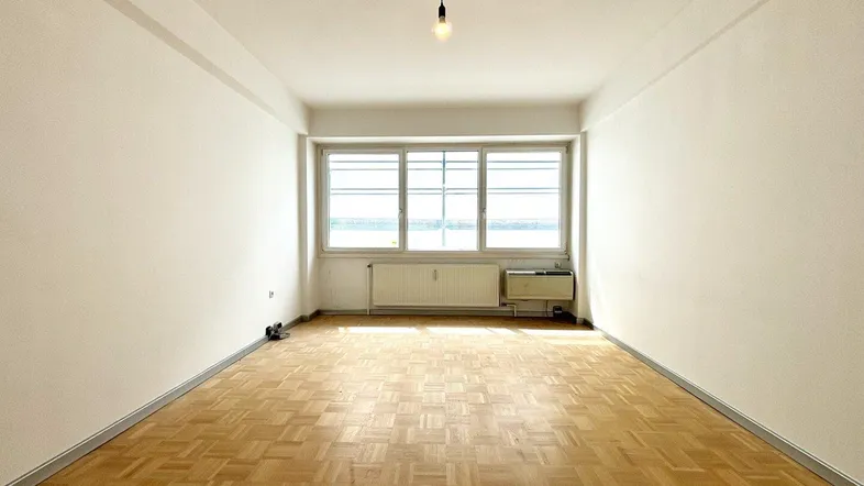 Heller, leerer Raum mit Parkettboden und großen Fenstern, ideal als Wohnzimmer nutzbar.