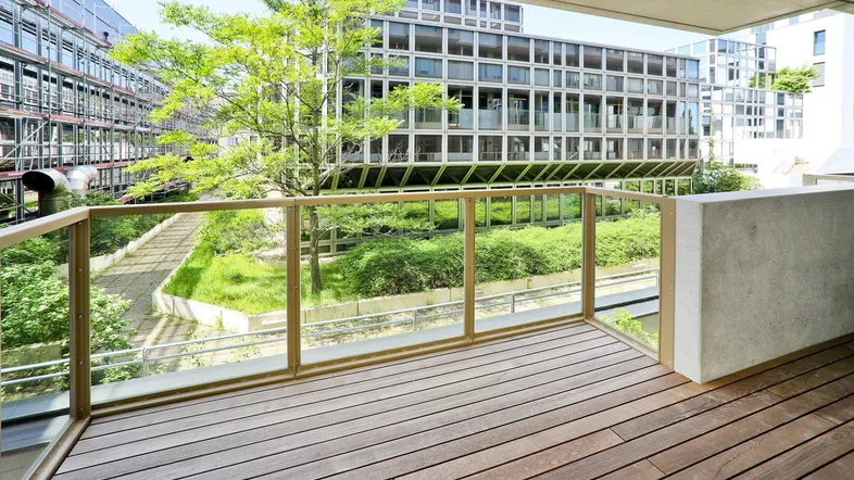 Geräumiger Balkon mit Holzboden und Glasgeländer, bietet Blick auf den begrünten Innenhof und moderne Gebäude.