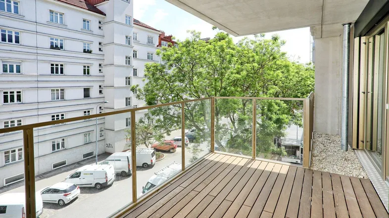 Moderner Balkon mit Holzfußboden und Glasgeländer, bietet Blick auf die umliegenden Gebäude und Bäume.