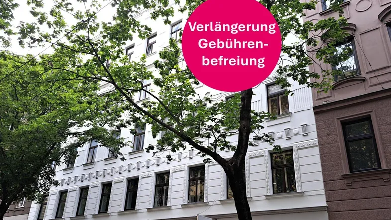 Elegante weiße Altbaufassade mit grünen Bäumen davor, daneben ein braunes Gebäude, teilweise von einem Werbehinweis verdeckt.