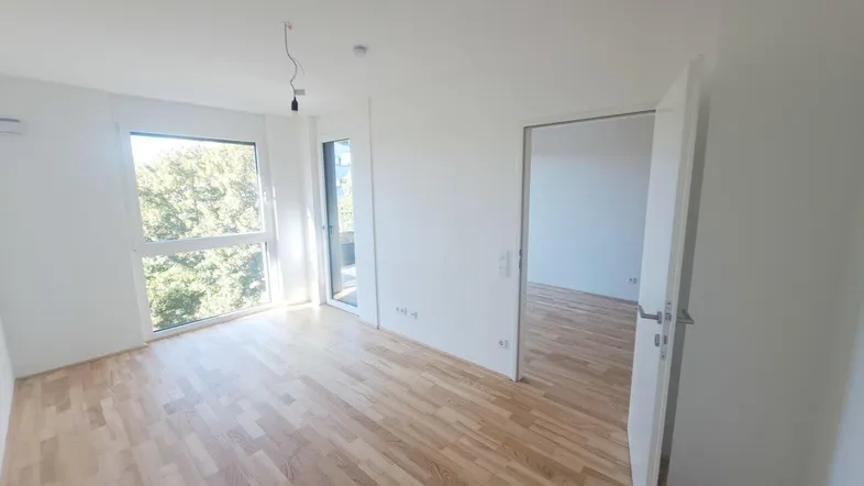 Helles Zimmer mit Holzboden und großen Fenstern, die viel Tageslicht hereinlassen.