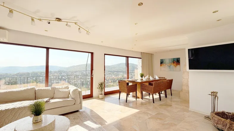 Helles Wohn- und Esszimmer mit großen Fenstern, Fliesenboden und weitem Blick über die grüne Landschaft.