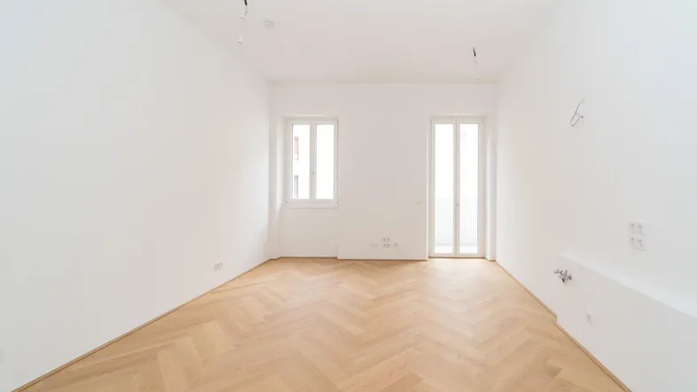Heller, leerer Raum mit Fischgrätparkettboden, Fenster und Zugang zu einem Außenbereich.