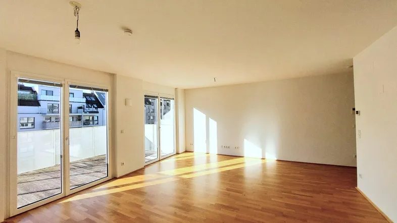 Helles, leeres Wohnzimmer mit Parkettboden und Zugang zu einem Balkon, durchflutet von Sonnenlicht.